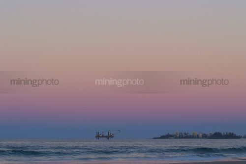 Offshore desalination plant drill rig working close to shore with headland and residents cloes by.  beach and waves in foreground. shot at sunset, good colours. - Mining Photo Stock Library