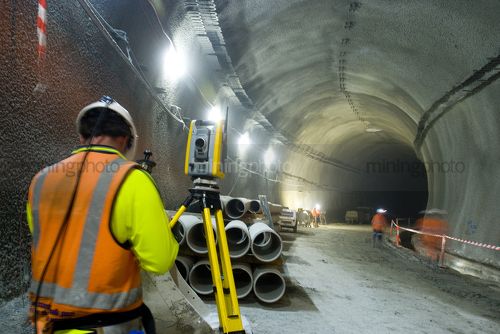 Underground civil road tunnel surveyor working on road construction in the tunnel.  shot from behind.  other workers blurred movement through the shot. - Mining Photo Stock Library