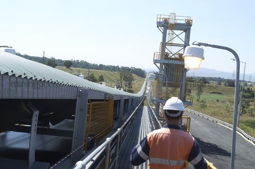 Mine worker in full PPE on walkway next to moving covered overland coal conveyor.  overland conveyor stretched to horizon. - Mining Photo Stock Library