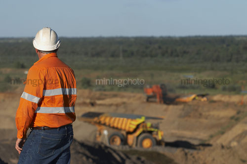 Mine worker in full PPE observing haul truck and mine workings at open cut mine. - Mining Photo Stock Library