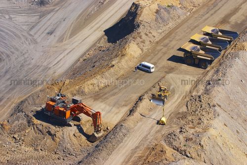 Large excavator diggin overburden in open cut coal mine.  haul trucks parked up at go line in background.  aerial vertical image.  light vehicle mini bus transporting truck drivers in middle ground. - Mining Photo Stock Library