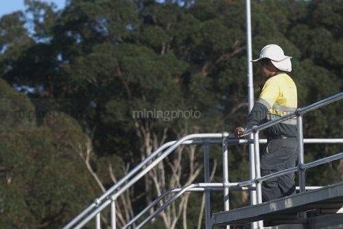 Worker in full PPE talking on radio while looking out over water treatment plant.  worker is at the top of walkway with one hand on railing. - Mining Photo Stock Library