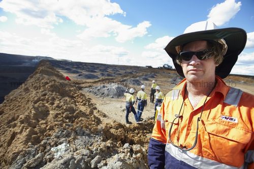 Mine site worker looking straight at the camera in open cut mine.  team workers pointing and working behind.  camera framed for double page spread. - Mining Photo Stock Library