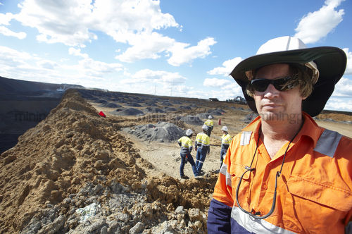 Mine site worker looking straight at the camera in open cut mine.  team workers pointing and working behind.  camera framed for double page spread. - Mining Photo Stock Library