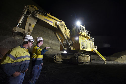 Shot at night, two 2 mine site workers in full PPE down in the open cut coal mine pit discussing next move with large excavator with lights on in the background. - Mining Photo Stock Library