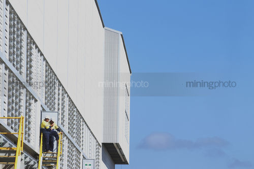 Two mine site workers in discussion standing on a  platform at the edge of shed platform.  blue sky behind. - Mining Photo Stock Library