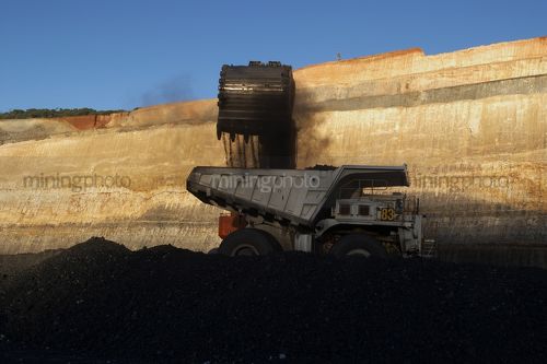 Generic photo of digger bucket loading coal into haul truck in open cut mine.  high walls and coal seam and blue sky in background. - Mining Photo Stock Library