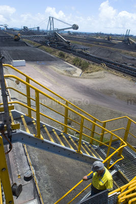 Mine site worker in full PPE walking down stairs with 3 three points of contact.  coal terminal with reclaimers in the background.  vertical photo. - Mining Photo Stock Library