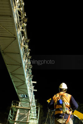 Rigger worker in full PPE including a harness working under a bridge at night.  crane and scaffolding in the background.  shot from behind. - Mining Photo Stock Library