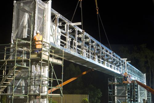 Workers in full PPE installing a gantry bridge over a motorway during the evening.  Scaffolding installed on both sides of the bridge. - Mining Photo Stock Library