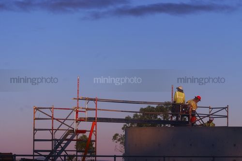 Workers in full PPE walking into confined space area from scaffolding platform.  shot during dawn light. - Mining Photo Stock Library
