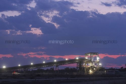 Night time photo of a covered coal conveyor into a hopper. light vehicle with lights on nearby for scale. - Mining Photo Stock Library