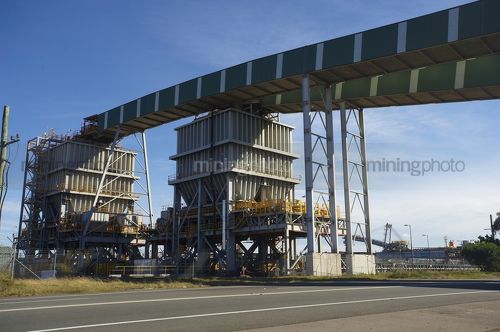 Enclosed coal conveyor crossing high over a road.  Ship loader in the background. - Mining Photo Stock Library