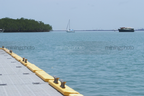 Looking over edge of a marine jetty with boats and mangroves on the horizon line. - Mining Photo Stock Library