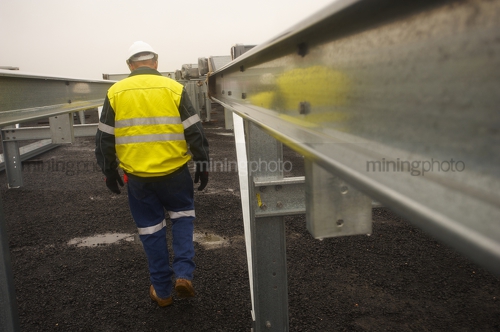 Construction manager in full yellow PPE walking amongst construction steel work.  steel is up close in foreground. - Mining Photo Stock Library