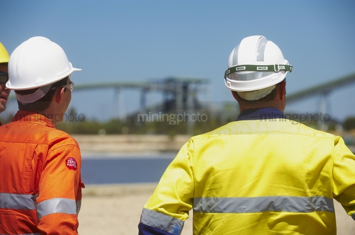 Two  mine site construction workers looking across a site.  large conveyor and stock pile in background. - Mining Photo Stock Library