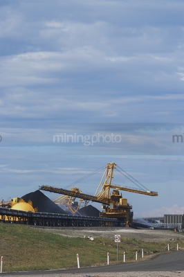 Photo of large shiploader and reclaimer working on coal stockpiles. light vehicle sealed road in foreground.  vertical photo. - Mining Photo Stock Library