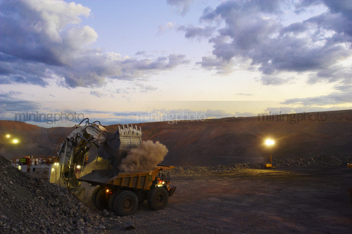 Great dusk photo of a digger loading a haul truck at open cut mine site.  lights are on. - Mining Photo Stock Library