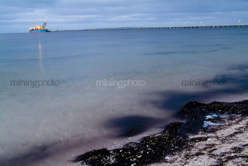 Ship and very long wharf in the distance being loaded. taken from the beach at water level.  - Mining Photo Stock Library