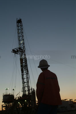 Silhouette of a drill rig worker with the derrick behind at dusk. - Mining Photo Stock Library
