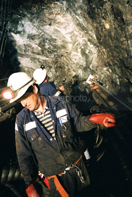 Underground worker returning to machinery with other miner inspecting roof in background. - Mining Photo Stock Library