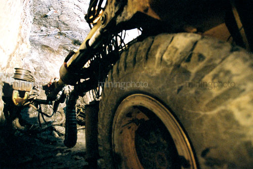 Underground jumbo machinery drilling holes for mesh anchors.  shot looking along from back wheel. - Mining Photo Stock Library