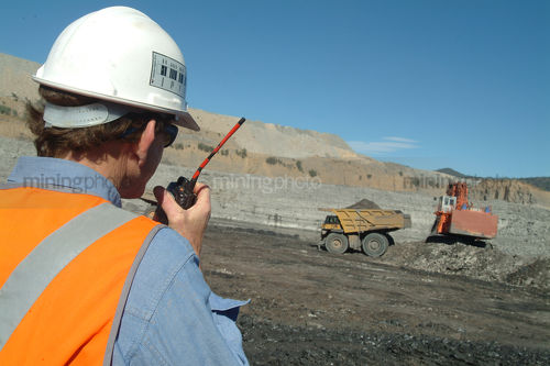 Mine site supervisor in full ppe talking into radio giving instructions to truck and digger.  shot from behind - Mining Photo Stock Library