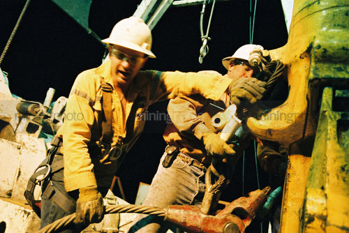 Great teamwork photo of two oil rig workers wrestle a huge cable onto the hook of the derrick. shot at night. - Mining Photo Stock Library