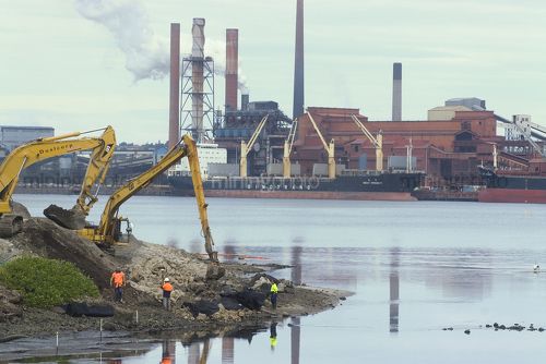 Long arm reach excavator working on edge of port with workers supervising. ship terminal and wharf in background. - Mining Photo Stock Library