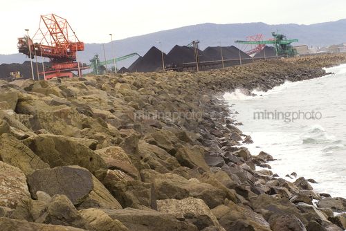 Reclaimers stockpiling coal at terminal with ocean surf pounding rock retaining wall in foreground.  shot from rock wall. - Mining Photo Stock Library