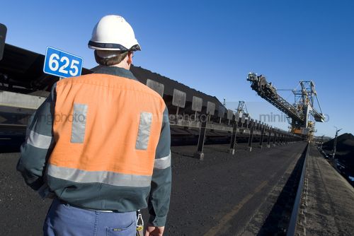 Mine worker in ppe walking towards coal ship loader and reclaimer.  shot from behind. - Mining Photo Stock Library