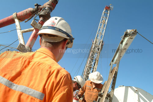 Oil and gas rig workers working together on a rig with the derrick in the background. shot from behind. - Mining Photo Stock Library