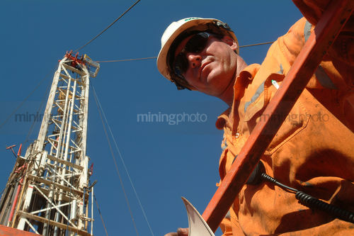 Oil and gas worker looking down into the camera from a walkway with the derrick behind - Mining Photo Stock Library