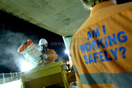 Construction worker wearing a safety mask works with concrete bags at night. another worker with signage on his shirt promoting safe work practices is in foreground. - Mining Photo Stock Library