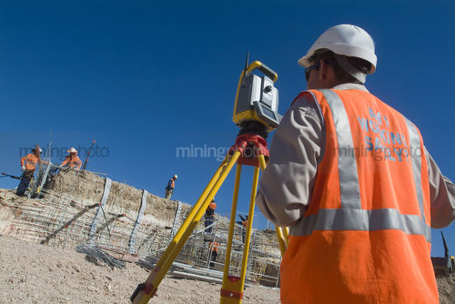 Surveyor on mine site with instrument and construction workers on steel form work in background. shot from behind to see instrument and workers. - Mining Photo Stock Library