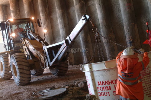 Construction worker in a tunnel putting large steel manhole into place with a loader and chain. - Mining Photo Stock Library