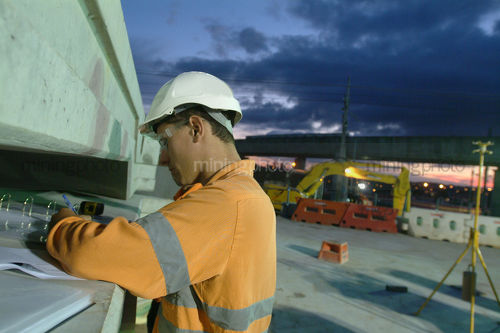 Site worker checks over infrastructure plans at dawn on work site. crane and mobile lights in the background. - Mining Photo Stock Library