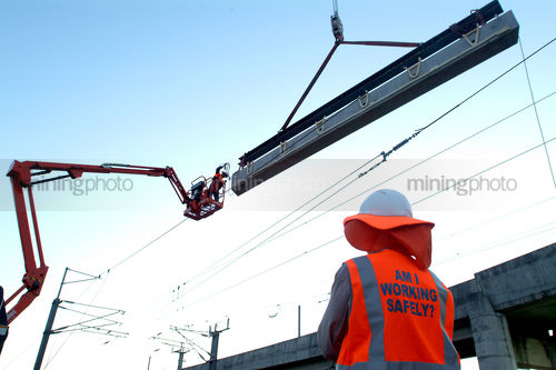 Site worker watching large concrete girder being craned into position with overhead rail electricity wires. - Mining Photo Stock Library