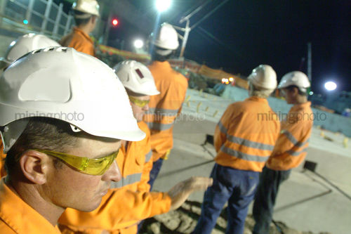 Close up of construction worker watching works with group of workers in background. shot at night under site lights. great recruitment shot - Mining Photo Stock Library