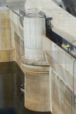 Worker in ppe walking along the top of a dam with the wall and water in foreground.  aerial and vertical shot. - Mining Photo Stock Library