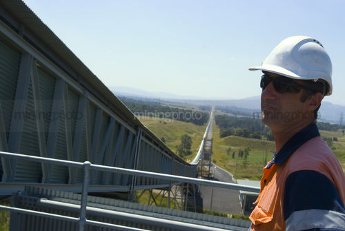 Male engineer in hard hat up close on tower with covered conveyor sweeping away into the background for many kilometres. - Mining Photo Stock Library