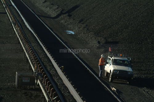Worker in ppe and his mine ready light vehicle next to coal loader tracks and stockpiled coal - Mining Photo Stock Library