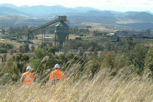 Two mine workers in PPE walk down hill at wash plant. distant shot of coal wash plant with rail and mountains in background.  good revegetation example in foreground. - Mining Photo Stock Library