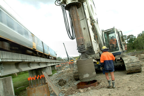 Worker drilling pylons  net to light Rail infrastructure track duplication project. train passing by.  great shot. - Mining Photo Stock Library