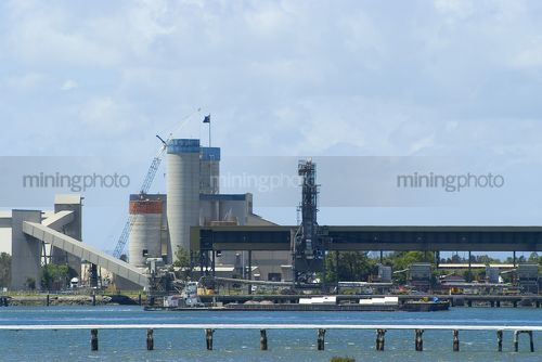 Large concrrete factory based on river bank with a tugboat moving a barge in foreground. industry - Mining Photo Stock Library