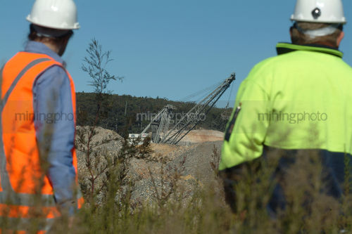 Two mine workers in PPE standing amongst revegetation with dragline working in open cut coal mine pit behind. - Mining Photo Stock Library