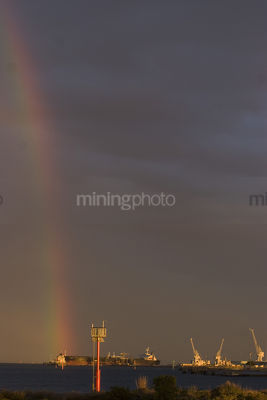 Rainbow sunset over ocean with shipping signal pylon in foreground and ships being loaded in the background.  vertical shot. - Mining Photo Stock Library