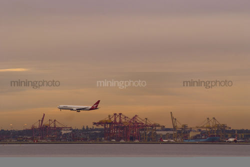 Plane about to land at airport with shipping port and wharf loaders underneath and in background. great colours at sunset.  very clean shot, good panorama strip. - Mining Photo Stock Library