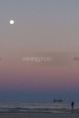 Fisherman on the beach with offshore desalination plant drill rig working close to shore. headland and close by.  moon rising. shot at sunset, good colours. - Mining Photo Stock Library