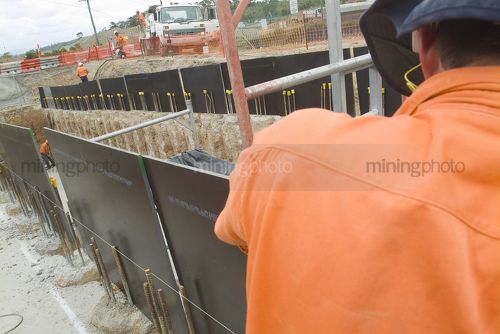 Workers on construction infrastructure site forming up boxing for concrete to be poured.  shot over the back of a worker in full PPE safety gear. - Mining Photo Stock Library